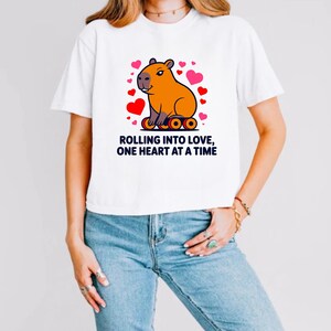 May include: White t-shirt featuring a cartoon capybara with a heart-shaped design. The capybara is orange and surrounded by red and pink hearts. The text "ROLLING INTO LOVE, ONE HEART AT A TIME" is printed below the graphic in black.