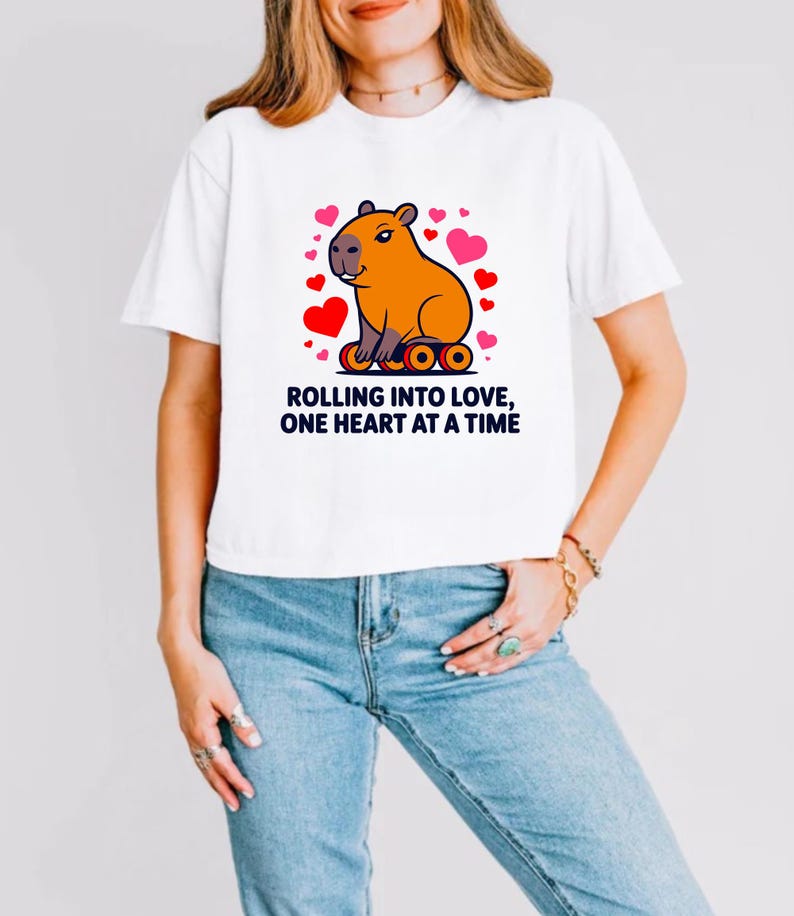 May include: White t-shirt featuring a cartoon capybara with a heart-shaped design. The capybara is orange and surrounded by red and pink hearts. The text "ROLLING INTO LOVE, ONE HEART AT A TIME" is printed below the graphic in black.