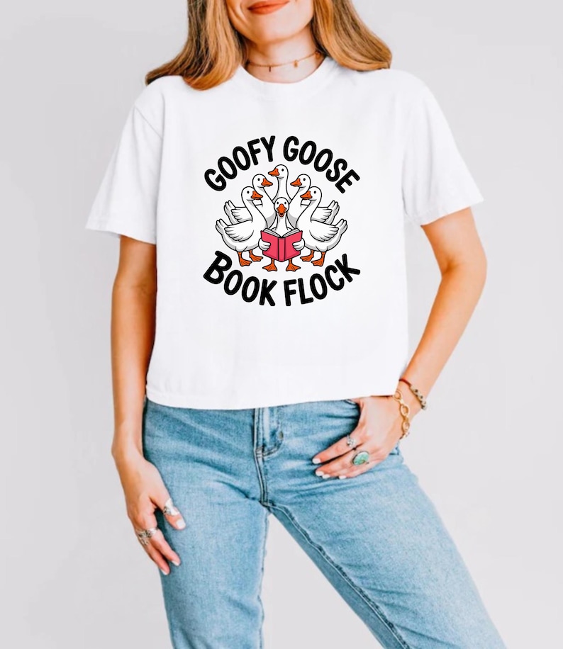 May include: White t-shirt with the text "GOOFY GOOSE BOOK FLOCK" in black, surrounding a graphic of geese reading a book. The geese are white with orange beaks and feet. The person is wearing blue jeans.