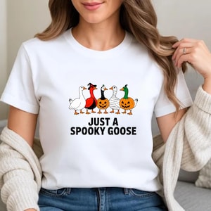 May include: White t-shirt with the text "JUST A SPOOKY GOOSE" in black. The shirt features a graphic of five cartoon geese dressed in Halloween costumes, including a witch hat, mummy wrap, and pumpkin.