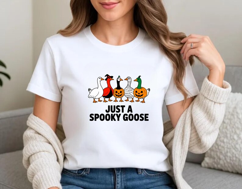 May include: White t-shirt with the text "JUST A SPOOKY GOOSE" in black. The shirt features a graphic of five cartoon geese dressed in Halloween costumes, including a witch hat, mummy wrap, and pumpkin.