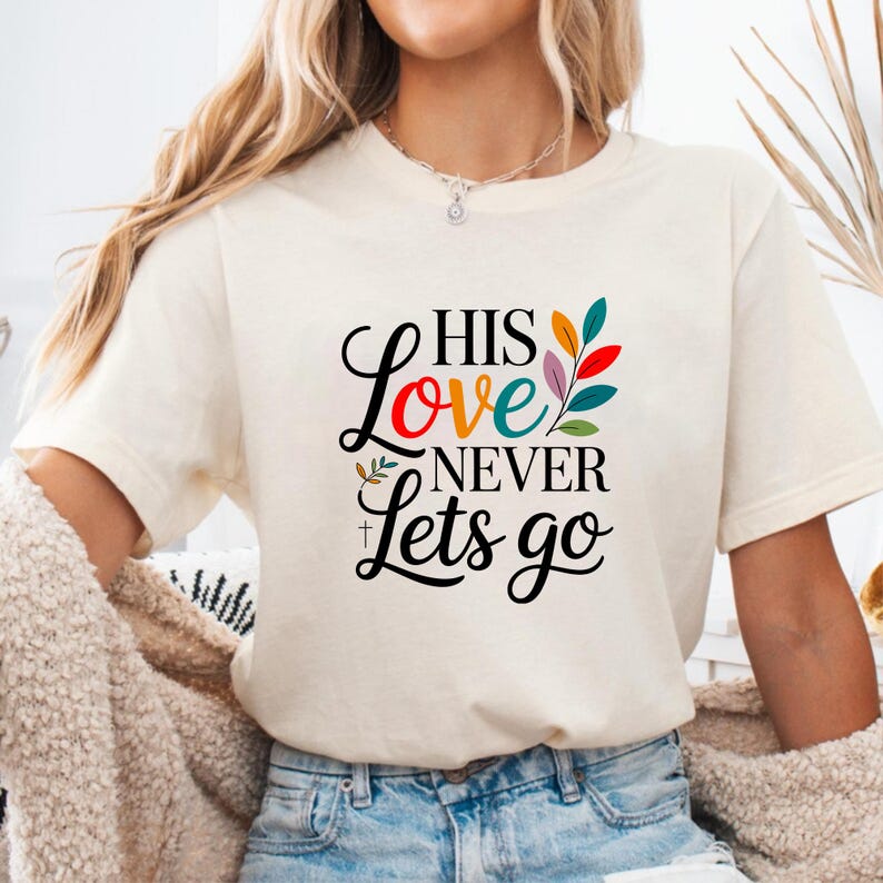 Op de afbeelding: Cr&egrave;mekleurig T-shirt met de tekst "HIS Love NEVER Lets go" in zwart script en kleurrijke accenten. Het woord "Love" is in rood, oranje en blauwgroen. Het shirt heeft een ronde hals en korte mouwen.