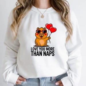 May include: White sweatshirt featuring a cartoon groundhog wearing heart-shaped sunglasses and holding two red heart balloons. The text "LOVE YOU MORE THAN NAPS" is printed below the groundhog in black letters.