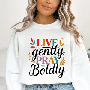 Op de afbeelding: Witte sweatshirt met de tekst "LIVE gently, PRAY Boldly" in kleurrijke letters. Het ontwerp bevat bloemen- en bladaccenten. De sweatshirt heeft een ronde hals.