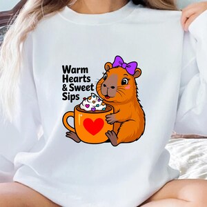 May include: White sweatshirt featuring a cartoon capybara with a purple bow, holding an orange mug with a heart and whipped cream. The text "Warm Hearts & Sweet Sips" is printed on the shirt. A cozy and cute design.