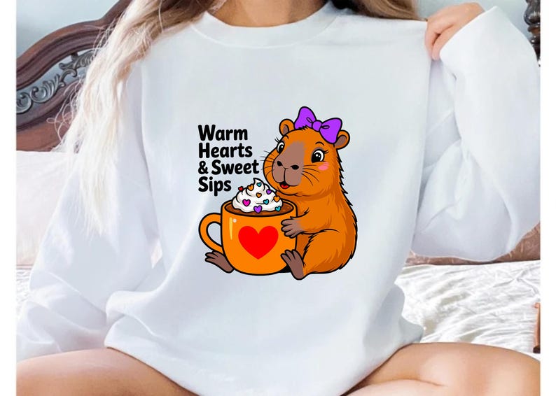 May include: White sweatshirt featuring a cartoon capybara with a purple bow, holding an orange mug with a heart and whipped cream. The text "Warm Hearts & Sweet Sips" is printed on the shirt. A cozy and cute design.