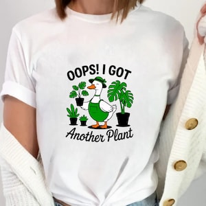May include: White t-shirt featuring a cartoon duck wearing a green hat and overalls, surrounded by potted plants. The text "OOPS! I GOT Another Plant" is printed below the duck. The shirt is a casual, comfortable style.