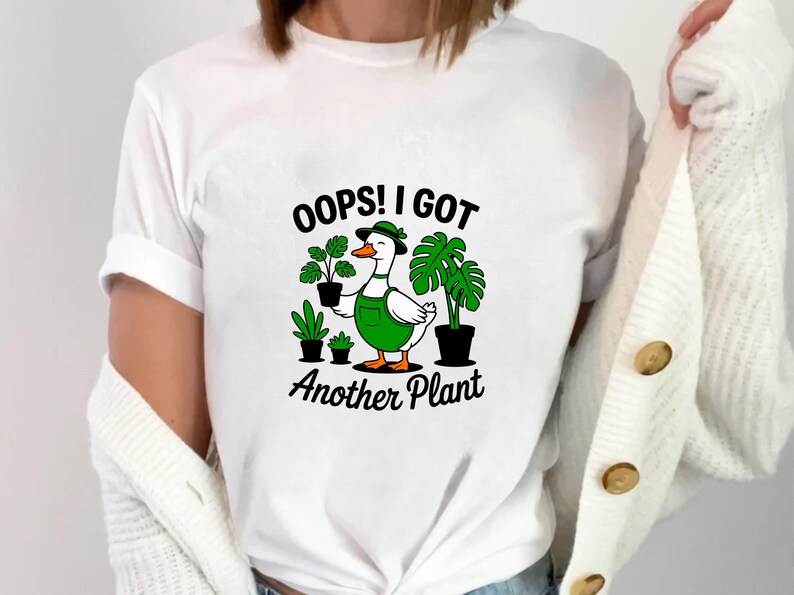 May include: White t-shirt featuring a cartoon duck wearing a green hat and overalls, surrounded by potted plants. The text "OOPS! I GOT Another Plant" is printed below the duck. The shirt is a casual, comfortable style.