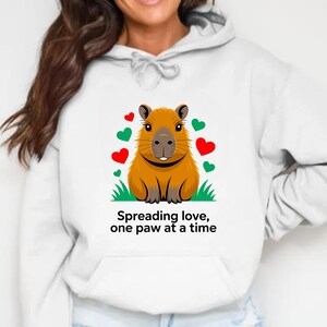 May include: White hoodie featuring a cartoon capybara with a smiling face, surrounded by red and green hearts. The text "Spreading love, one paw at a time" is printed below the capybara. The hoodie has a front pocket and a drawstring hood.