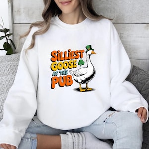 May include: White sweatshirt featuring a cartoon goose wearing a green top hat and a shamrock. The text reads "Silliest Goose at the Pub" in orange and yellow. The goose is black and white with yellow feet and beak.