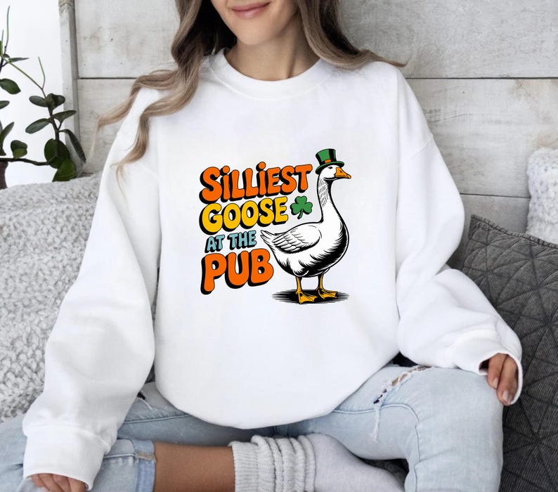 May include: White sweatshirt featuring a cartoon goose wearing a green top hat and a shamrock. The text reads "Silliest Goose at the Pub" in orange and yellow. The goose is black and white with yellow feet and beak.