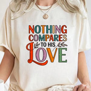 Op de afbeelding: Cr&egrave;mekleurig T-shirt met de tekst "NOTHING COMPARES TO HIS LOVE" in kleurrijke, retro letters. Het ontwerp bevat oranje, groene, roze en paarse tinten, met decoratieve bladaccenten. Een gouden ketting is zichtbaar.