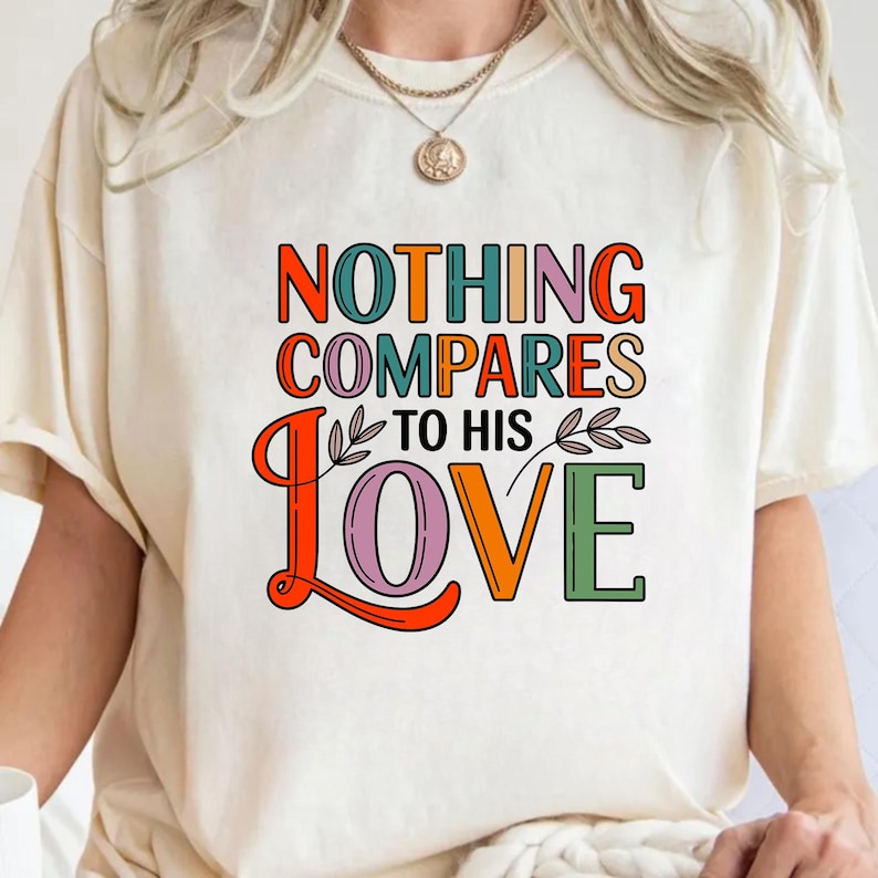 Op de afbeelding: Cr&egrave;mekleurig T-shirt met de tekst "NOTHING COMPARES TO HIS LOVE" in kleurrijke, retro letters. Het ontwerp bevat oranje, groene, roze en paarse tinten, met decoratieve bladaccenten. Een gouden ketting is zichtbaar.