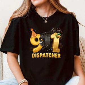 Puede incluir: Camiseta negra con un diseño gráfico dorado para un operador del 911. El diseño incluye los números "911", con un auricular integrado en el "1". La palabra "DISPATCHER" está impresa debajo en dorado.