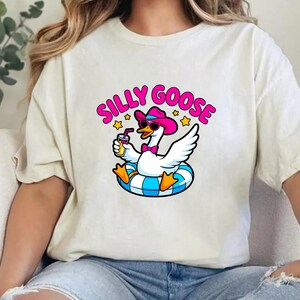 May include: Cream-colored t-shirt featuring a cartoon goose wearing sunglasses and a pink cowboy hat, floating on a blue and white inner tube. The text "SILLY GOOSE" is above the goose in pink and yellow.