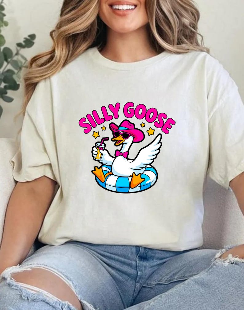 May include: Cream-colored t-shirt featuring a cartoon goose wearing sunglasses and a pink cowboy hat, floating on a blue and white inner tube. The text "SILLY GOOSE" is above the goose in pink and yellow.