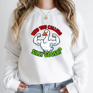 May include: White sweatshirt featuring a cartoon goose flexing its muscles. The goose has a red and orange beak and is surrounded by the text "WHO YOU CALLING SILLY GOOSE?" in red and green lettering. A fun, casual apparel item.