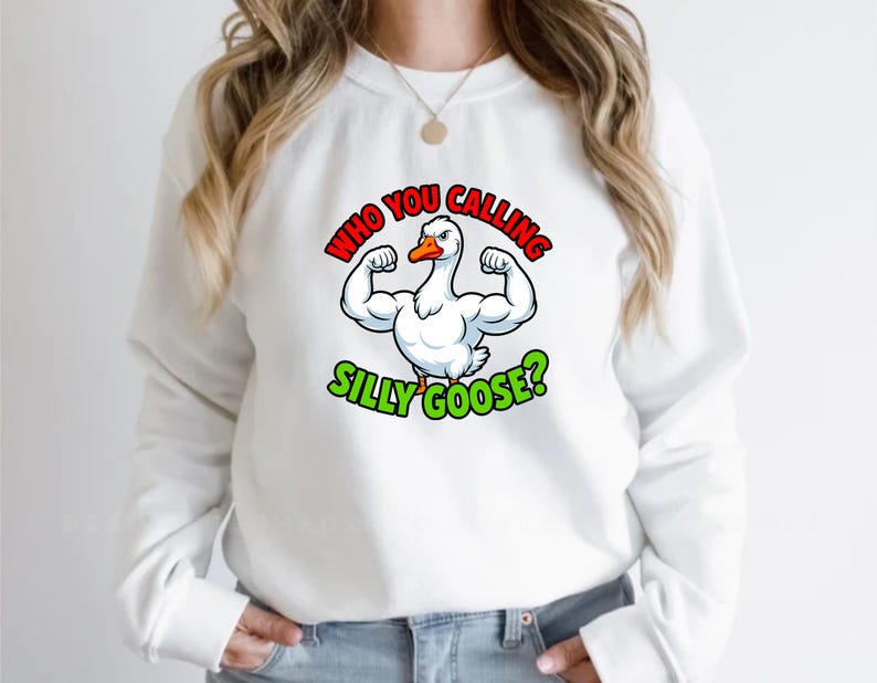 May include: White sweatshirt featuring a cartoon goose flexing its muscles. The goose has a red and orange beak and is surrounded by the text "WHO YOU CALLING SILLY GOOSE?" in red and green lettering. A fun, casual apparel item.