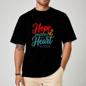Op de afbeelding: Zwart T-shirt met de tekst "Hope anchors my Heart in Him" in rode, turquoise en oranje letters. Een ankergrafiek is in het ontwerp opgenomen. Het shirt heeft korte mouwen en een ronde hals.
