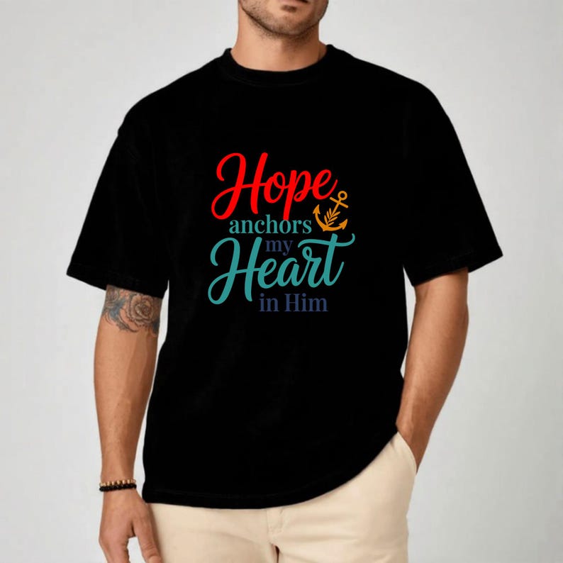 Op de afbeelding: Zwart T-shirt met de tekst "Hope anchors my Heart in Him" in rode, turquoise en oranje letters. Een ankergrafiek is in het ontwerp opgenomen. Het shirt heeft korte mouwen en een ronde hals.