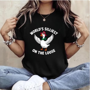 May include: Black t-shirt featuring a cartoon goose with outstretched wings, wearing a red hat and green scarf. The text "WORLD'S SILLIEST ON THE LOOSE" is printed above and below the goose. The shirt is worn by a person.