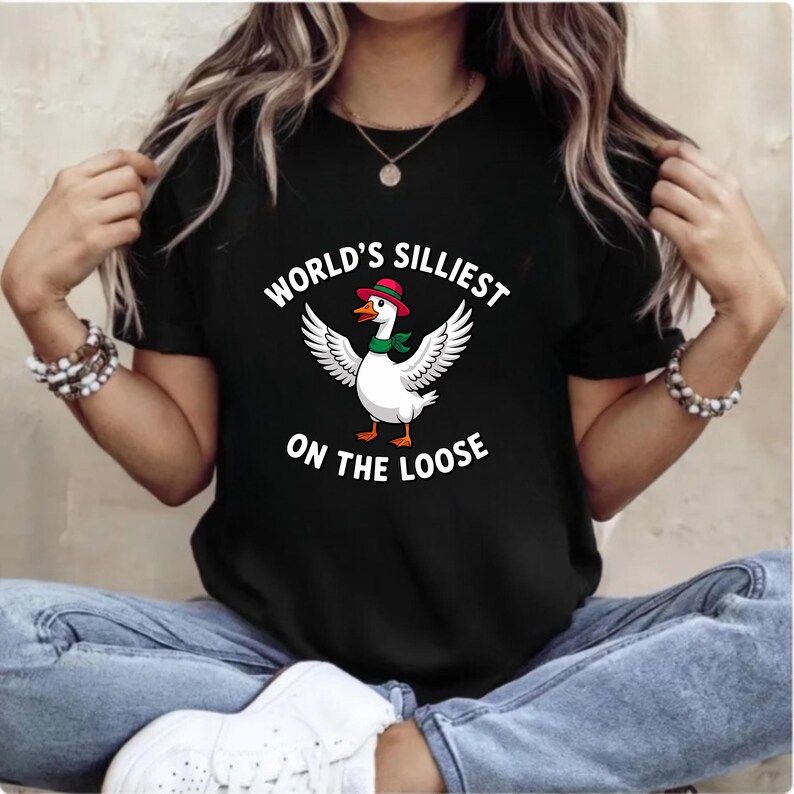 May include: Black t-shirt featuring a cartoon goose with outstretched wings, wearing a red hat and green scarf. The text "WORLD'S SILLIEST ON THE LOOSE" is printed above and below the goose. The shirt is worn by a person.