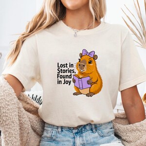 May include: Cream-colored t-shirt featuring a cartoon capybara reading a purple book and wearing a purple bow. The text "Lost in Stories, Found in Joy" is printed on the shirt in black. The shirt is made of a soft material.