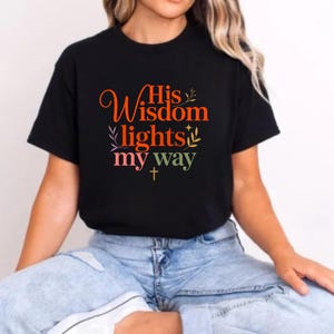 Op de afbeelding: Zwart T-shirt met de tekst "His Wisdom lights my way" in oranje, groen, roze en geel. Het ontwerp bevat een kruis en bloemaccenten. Het shirt heeft een ronde hals en korte mouwen.
