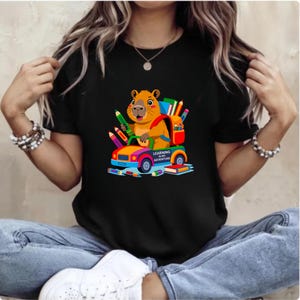May include: Black t-shirt featuring a colorful graphic of a cartoon beaver driving a car filled with pencils and books. The beaver has a backpack and the text "LEARNING IS MY ADVENTURE" is on the car. The shirt is a casual wear item.