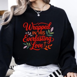 Op de afbeelding: Zwart sweatshirt met de tekst "Wrapped in His Everlasting Love" in rode letters. Het ontwerp bevat bloem- en bladaccenten in groen, roze en paars, evenals een klein kruis.