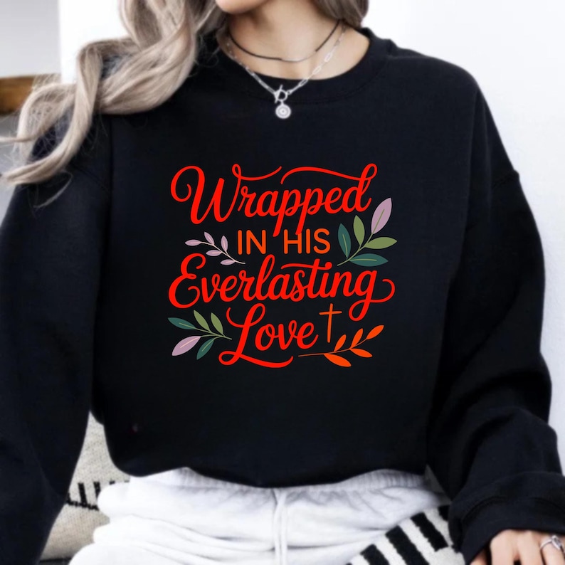 Op de afbeelding: Zwart sweatshirt met de tekst "Wrapped in His Everlasting Love" in rode letters. Het ontwerp bevat bloem- en bladaccenten in groen, roze en paars, evenals een klein kruis.