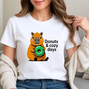 May include: White t-shirt featuring a cartoon capybara holding a green donut. The text "Donuts & cozy days" is printed on the shirt. The capybara is orange and brown with black accents. The donut is green with white sprinkles.