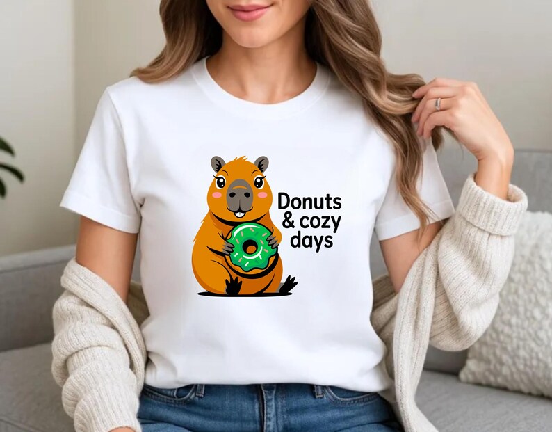 May include: White t-shirt featuring a cartoon capybara holding a green donut. The text "Donuts & cozy days" is printed on the shirt. The capybara is orange and brown with black accents. The donut is green with white sprinkles.