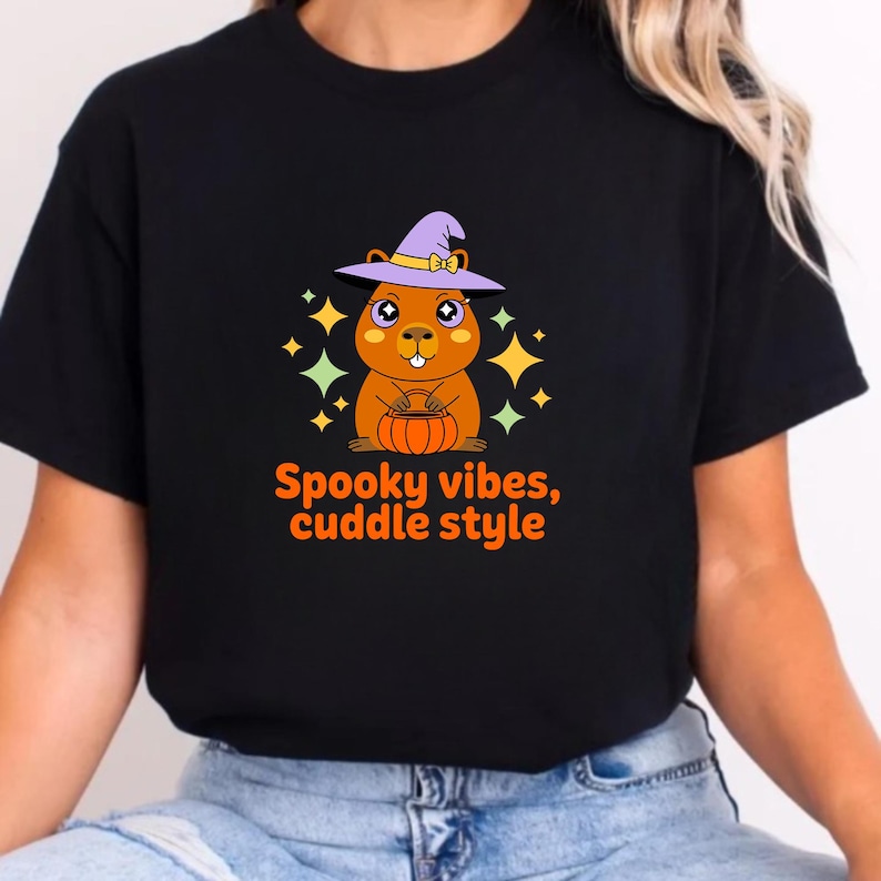 May include: Black t-shirt featuring a cartoon capybara wearing a purple witch hat and holding a pumpkin. The design includes the text "Spooky vibes, cuddle style" in orange. The background has yellow and orange star accents.