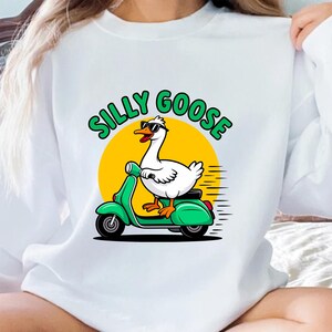May include: White sweatshirt featuring a cartoon goose wearing sunglasses and riding a green scooter. The text "SILLY GOOSE" is above the goose in green letters with a yellow sun background. A fun, whimsical design.