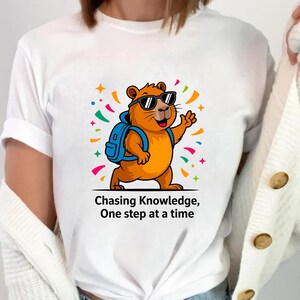 May include: White t-shirt featuring a cartoon groundhog wearing sunglasses and a backpack, waving with its right paw. The groundhog is orange with a blue backpack. The text "Chasing Knowledge, One step at a time" is below the image.