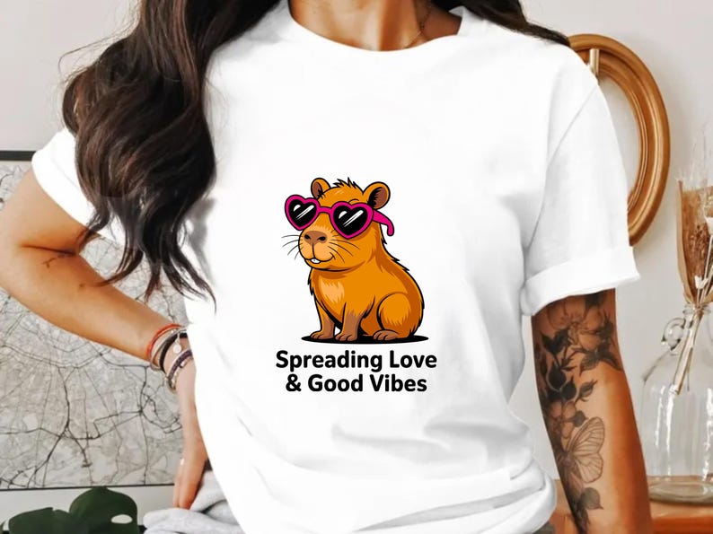May include: White t-shirt featuring a cartoon capybara wearing pink heart-shaped sunglasses. The capybara is orange and brown. Text below the graphic reads "Spreading Love & Good Vibes."