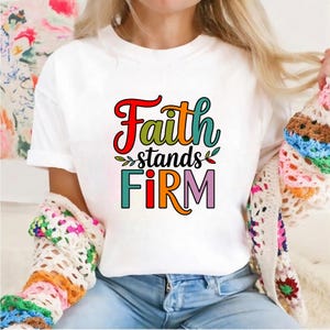 Op de afbeelding: Wit T-shirt met de tekst "Faith stands FIRM" in een kleurrijk, gestileerd lettertype. Het ontwerp bevat rode, groene, oranje en paarse letters. Een casual kledingstuk.