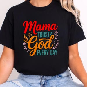 Op de afbeelding: Zwart t-shirt met de tekst "Mama Trusts God Every Day" in rood, oranje en turquoise. Het ontwerp bevat bloemmotieven en een kruis. Het t-shirt is gemaakt van zachte stof.
