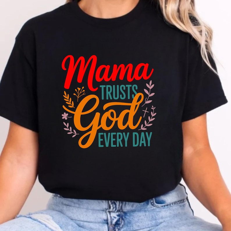Op de afbeelding: Zwart t-shirt met de tekst "Mama Trusts God Every Day" in rood, oranje en turquoise. Het ontwerp bevat bloemmotieven en een kruis. Het t-shirt is gemaakt van zachte stof.