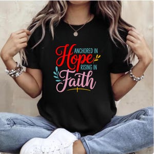 Op de afbeelding: Zwart T-shirt met de tekst "Anchored in Hope, Rising in Faith" in rode, roze en blauwe letters. Het ontwerp bevat bloemaccenten. De persoon draagt een blauwe spijkerbroek.