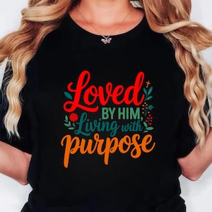 Op de afbeelding: Zwart T-shirt met de tekst "Loved by Him, Living with Purpose" in rode, groene en oranje letters. Het ontwerp bevat bloemaccenten en een klein kruis. Het shirt heeft een ronde hals.