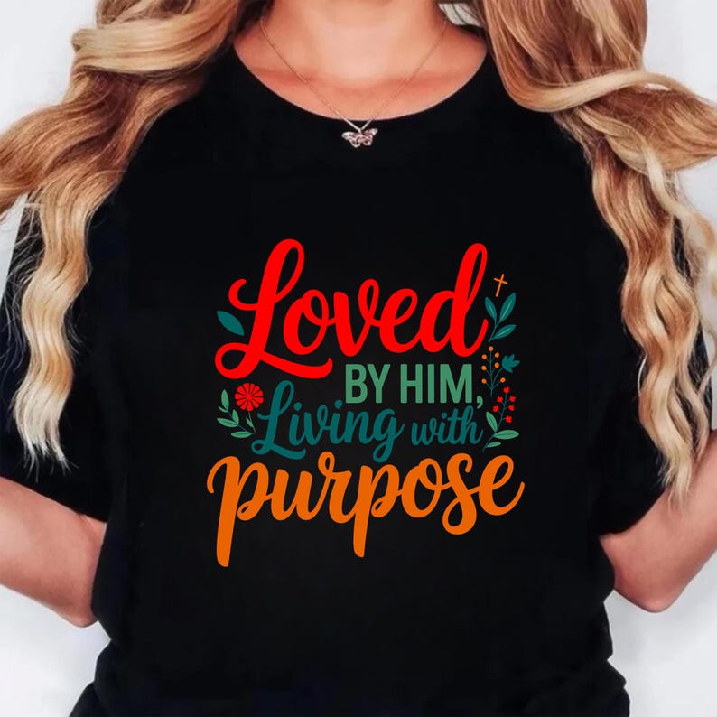 Op de afbeelding: Zwart T-shirt met de tekst "Loved by Him, Living with Purpose" in rode, groene en oranje letters. Het ontwerp bevat bloemaccenten en een klein kruis. Het shirt heeft een ronde hals.