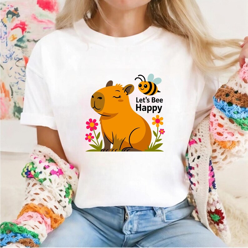 May include: White t-shirt featuring a cartoon capybara with closed eyes, surrounded by flowers and the text "Let's Bee Happy". A yellow and black bee is also present. The design is printed on a white t-shirt.