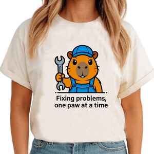 May include: Cream-colored t-shirt featuring a cartoon beaver wearing a blue cap and overalls, holding a wrench. The text "Fixing problems, one paw at a time" is printed below the beaver.