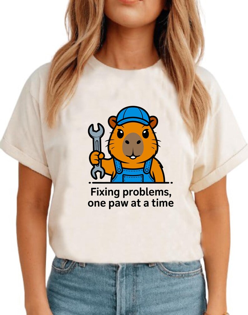 May include: Cream-colored t-shirt featuring a cartoon beaver wearing a blue cap and overalls, holding a wrench. The text "Fixing problems, one paw at a time" is printed below the beaver.