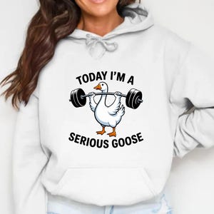May include: White hoodie featuring a cartoon goose lifting a barbell. The goose is white with orange feet and a yellow beak. Text on the hoodie reads "TODAY I'M A SERIOUS GOOSE".