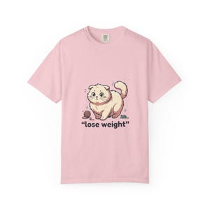 Könnte beinhalten: Hellrosa T-Shirt mit einer Cartoon-Katze und dem Text "lose weight". Die Katze ist beige, mit einem Wollknäuel und einer Maus. Das T-Shirt hat einen Rundhalsausschnitt und kurze Ärmel.