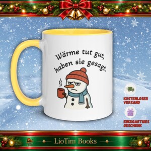 Könnte beinhalten: Ein weißer Keramikbecher mit gelbem Henkel und gelbem Innenrand. Der Becher zeigt eine Grafik eines mürrischen Schneemanns mit roter Mütze und blauem Schal, der eine rote Tasse hält. Über dem Schneemann steht der Text: Wärme tut gut, haben sie gesagt.