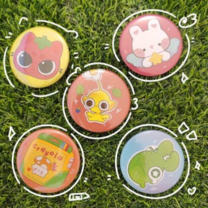 May include: Five colorful round button pins with cartoon designs. The pins feature a tomato-like character, a bunny holding a star, a yellow creature, a Crayola box with a mushroom, and a green dinosaur.